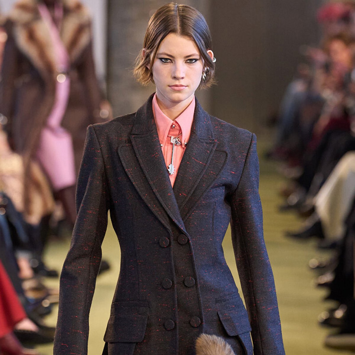 Model walking a fashion runway in a dark, double-breasted blazer over a pink shirt.