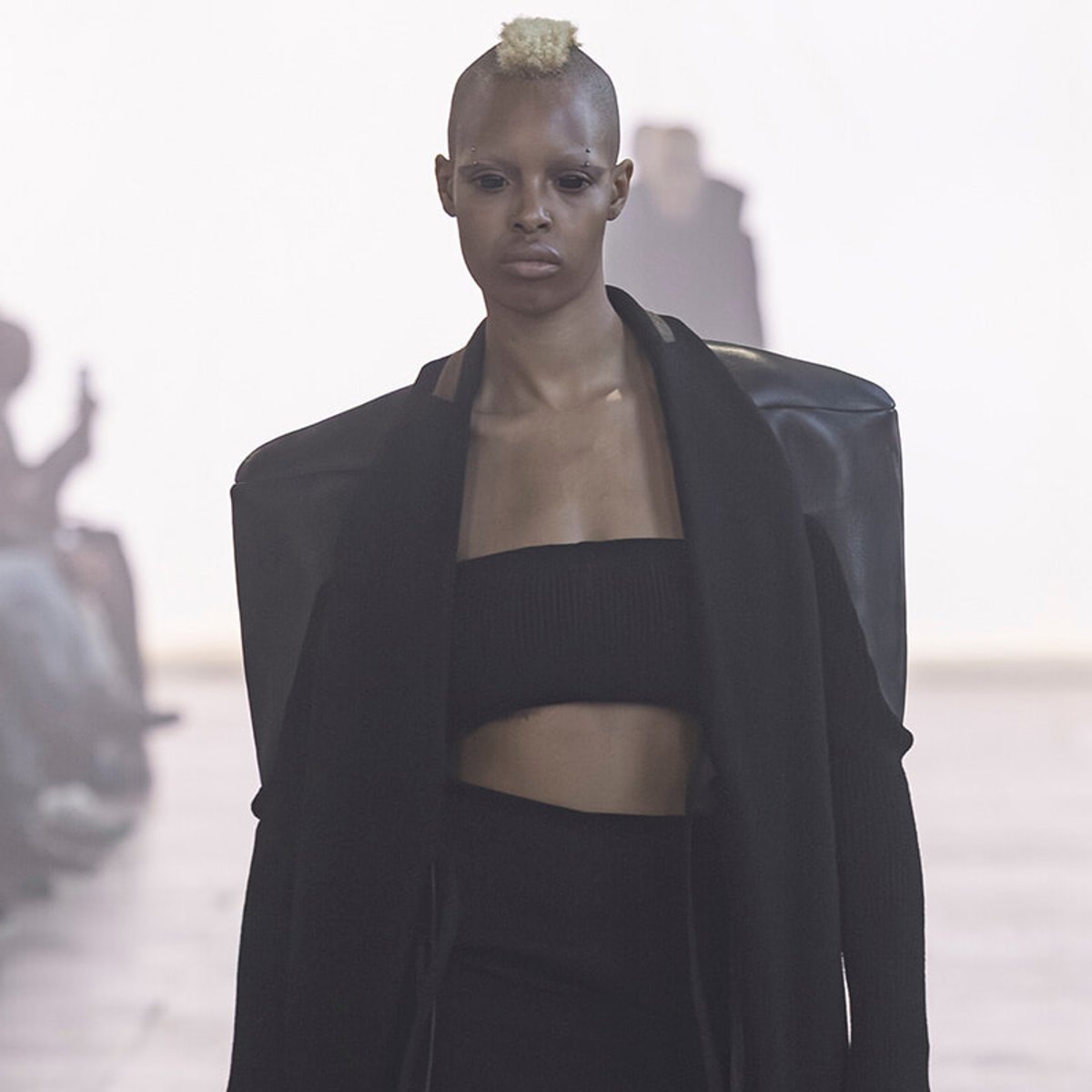 Model walks the runway in a black, oversized coat over a black bandeau top; pale blonde mohawk and serious expression dominate the look.