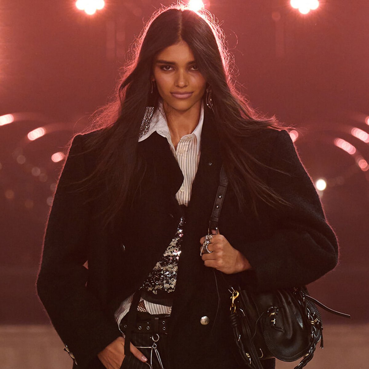 Model walking a runway in a black coat over a sequined top, white collared shirt, and a black handbag under red stage lights.