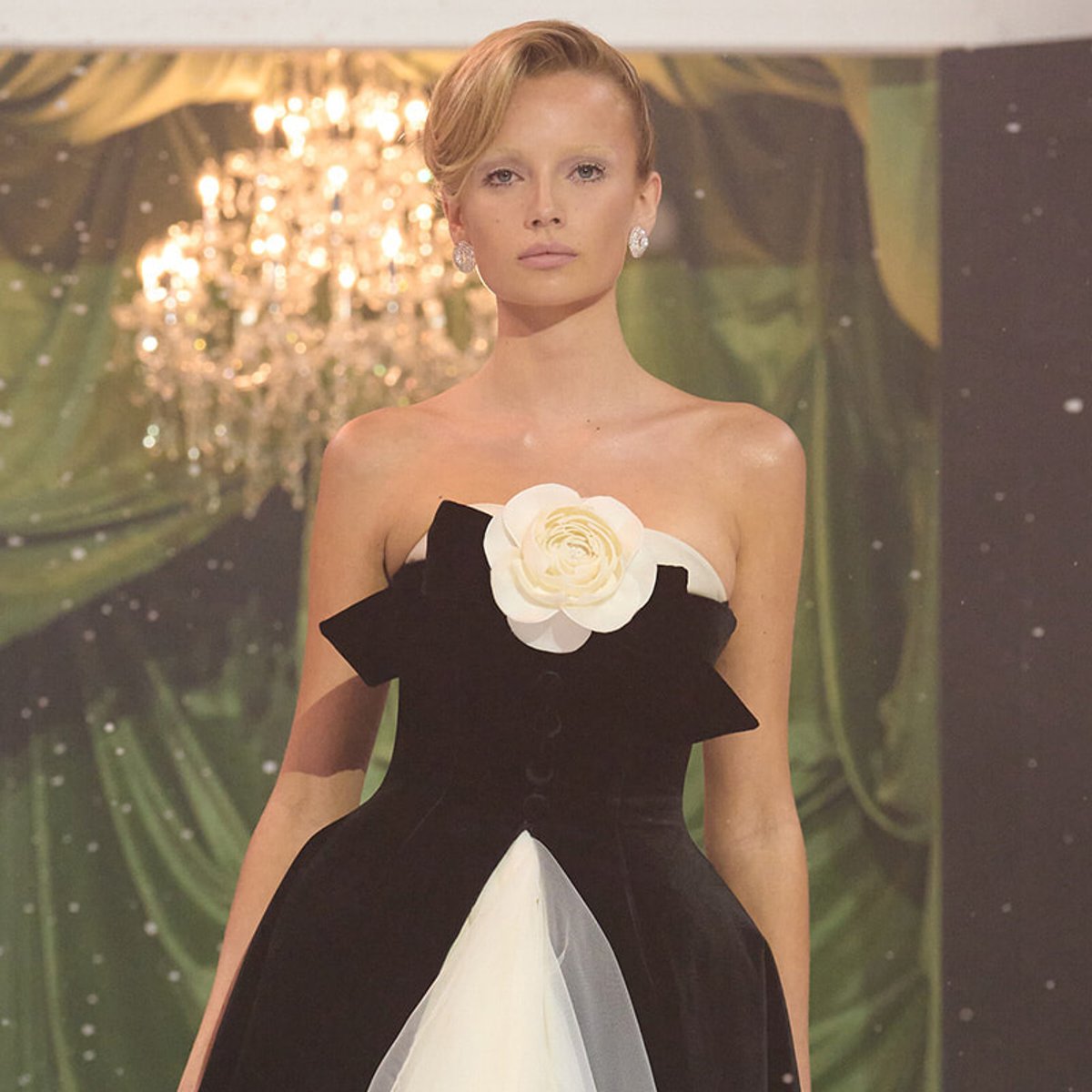 Runway model wearing a black strapless gown with a large white flower at the chest, chandelier backdrop behind.] ,