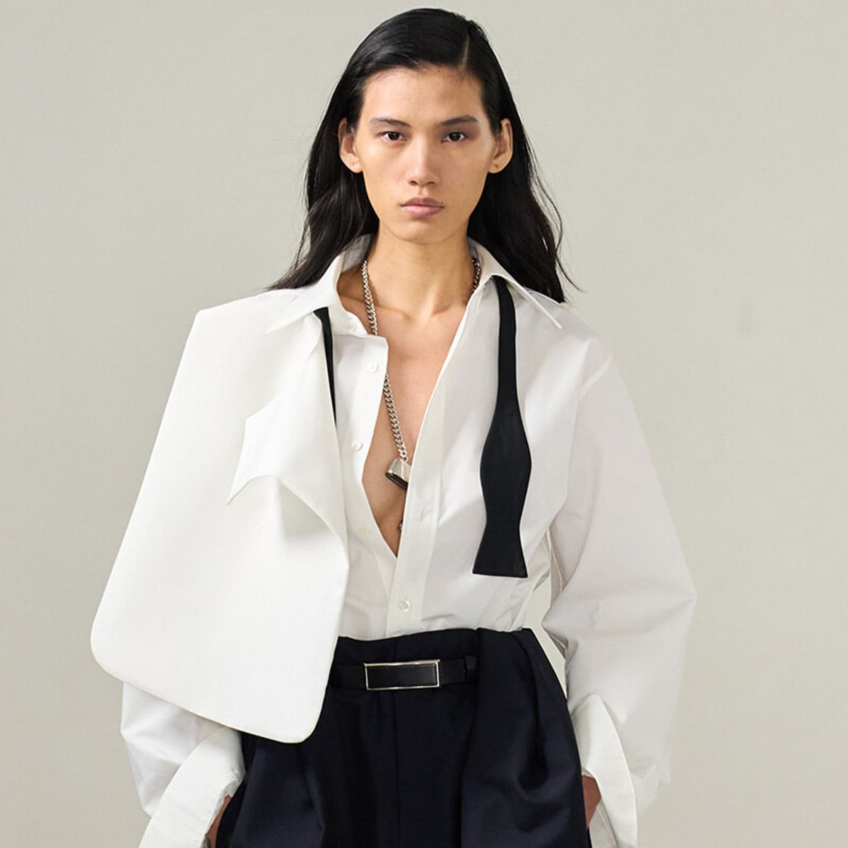 Fashion model in a crisp white oversized shirt with a black necktie detail, paired with high-waisted black trousers and a belt, posing against a neutral backdrop.
