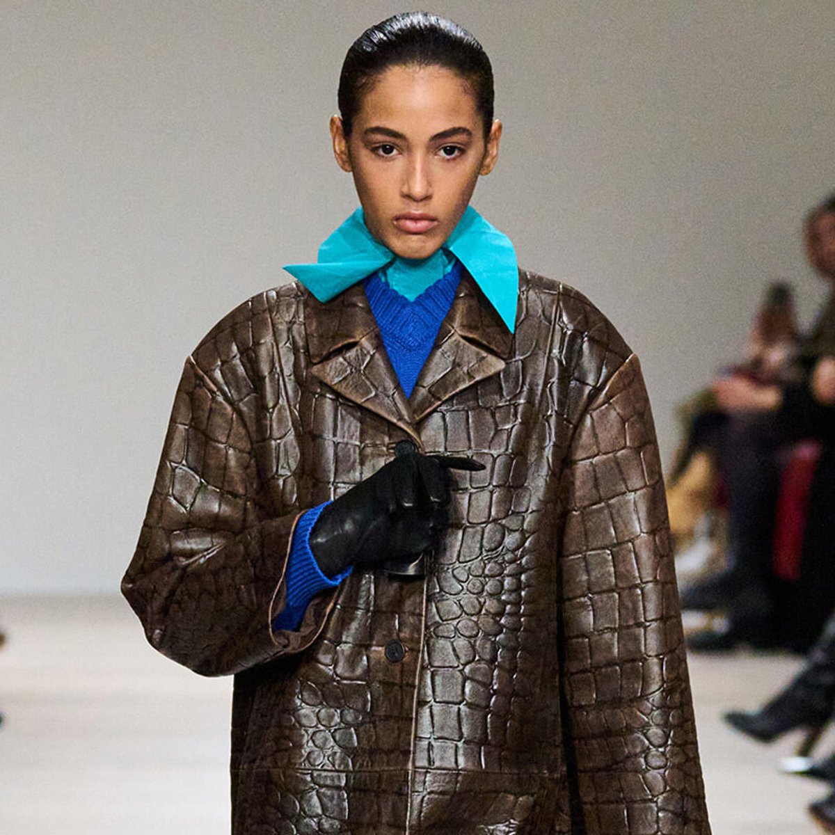 Model on a runway wearing a brown crocodile-textured coat over a blue sweater, with a teal oversized collar and black glove on one hand.