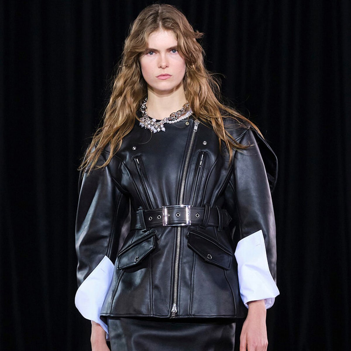 Fashion model wearing a black leather jacket with a belt, silver zippers, and layered pockets on a dark runway backdrop.