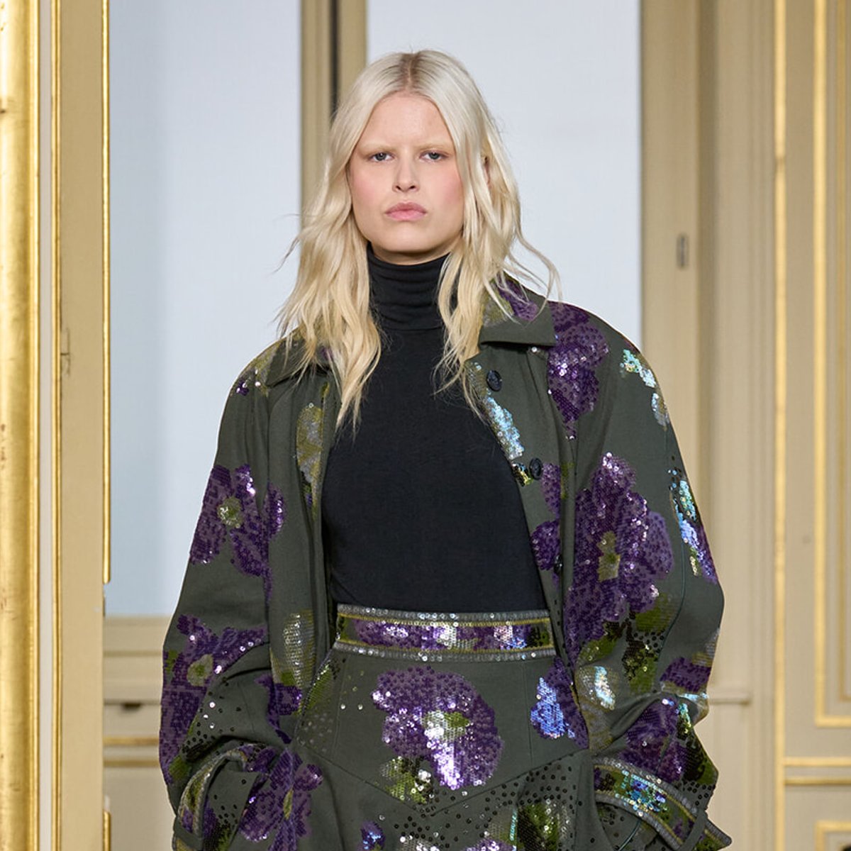 Model walking a runway in an oversized olive coat with purple sequins over a black turtleneck in a gilded, elegant setting.