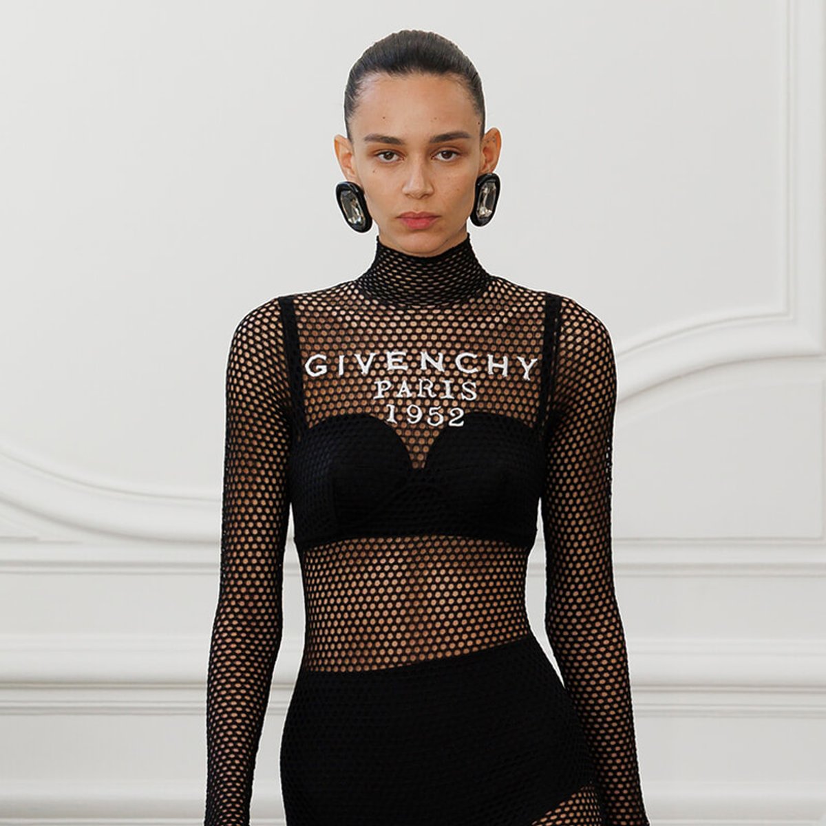 Model wearing a black fishnet mesh top with the words 'GIVENCHY PARIS 1952' across the chest, with large circular earrings in a white studio setting.