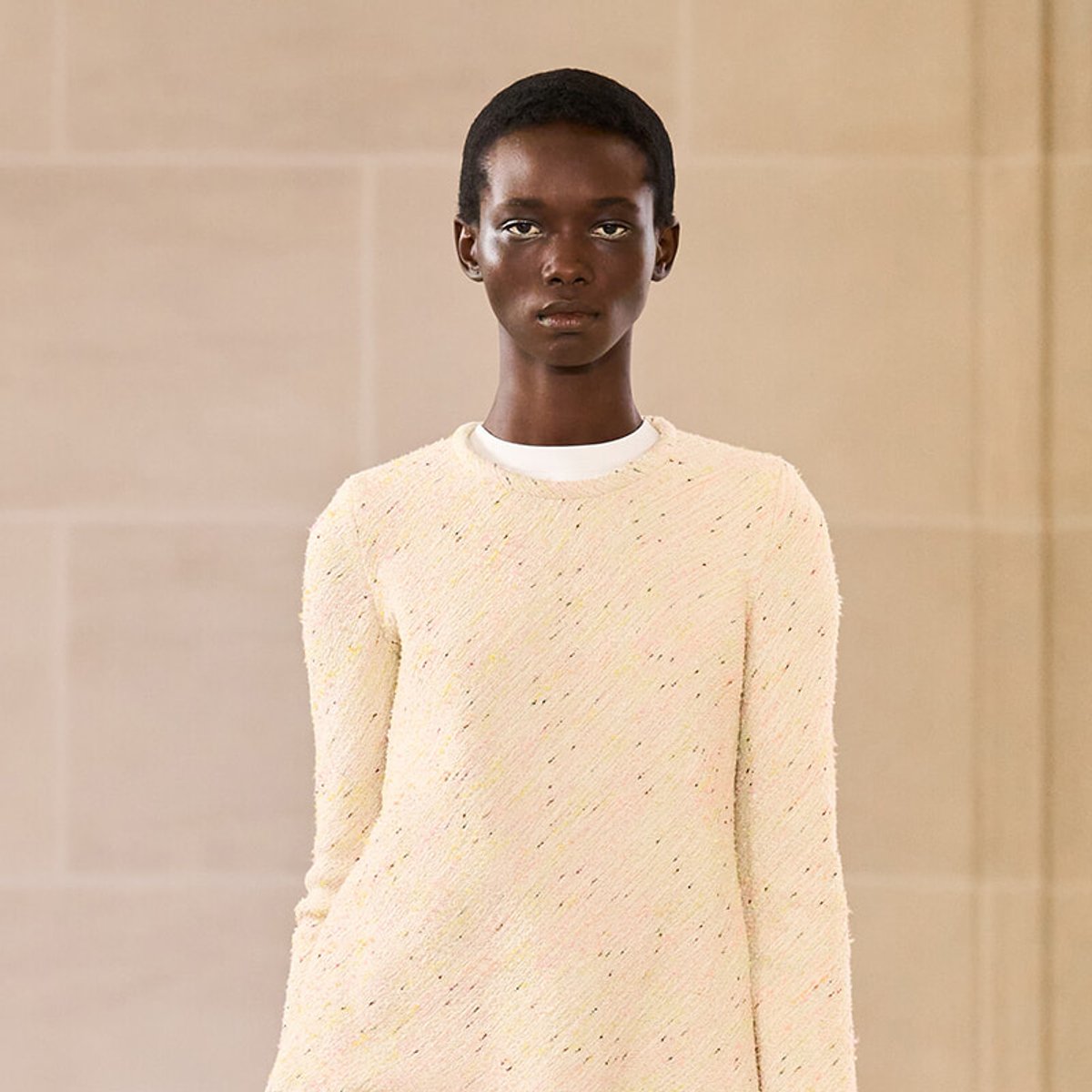 Model with dark skin wearing a cream textured knit sweater, standing against a beige wall backdrop.