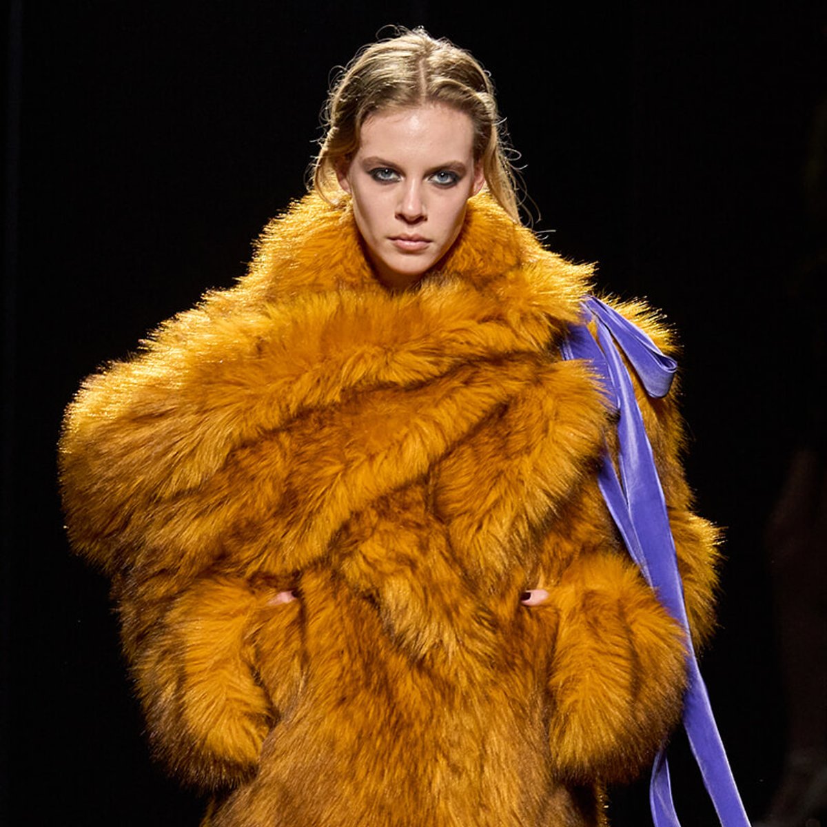 Model wearing a voluminous orange fur coat with a purple sash on a dark runway backdrop.