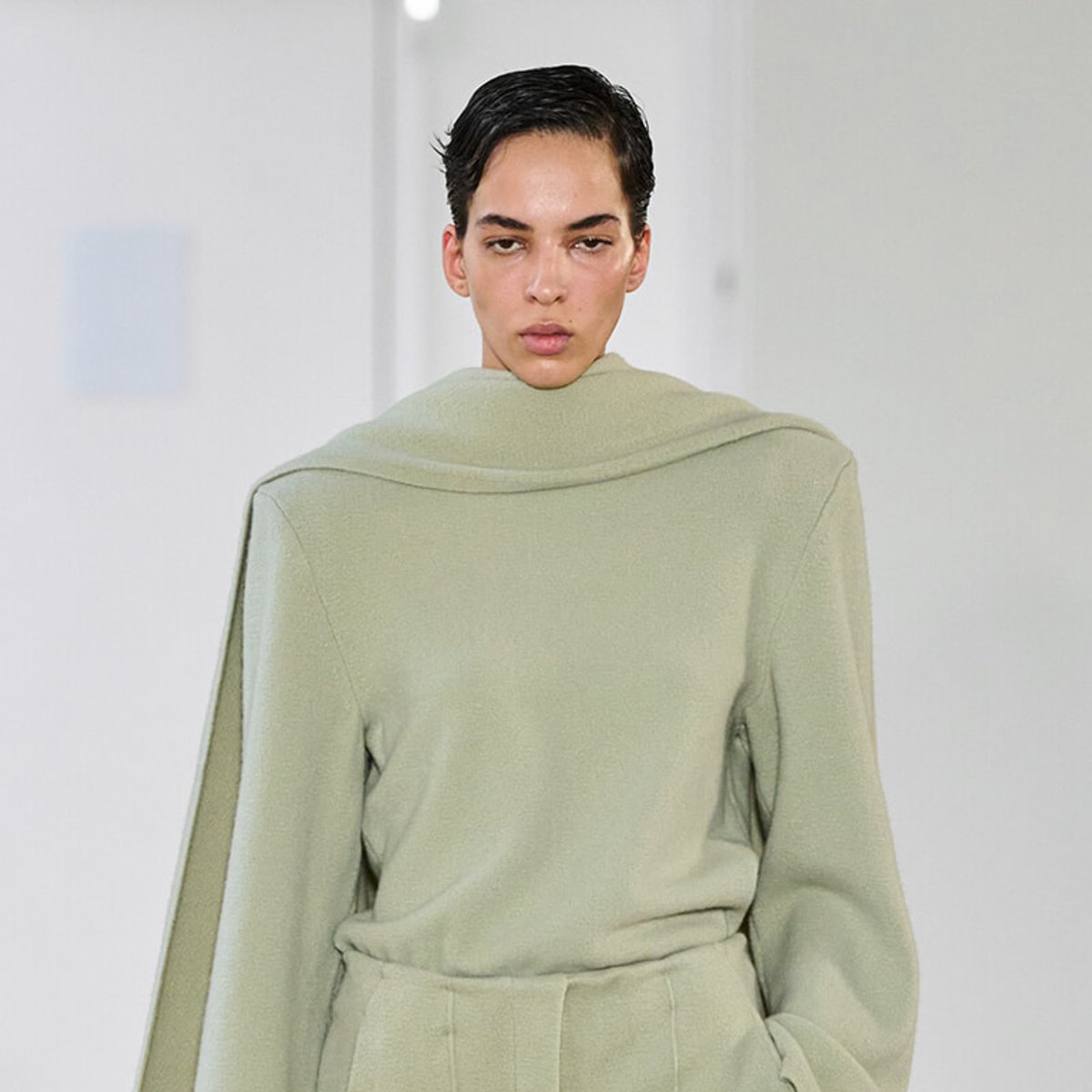 Model walking runway in a loose sage-green sweater with a high, draped collar and relaxed fit.