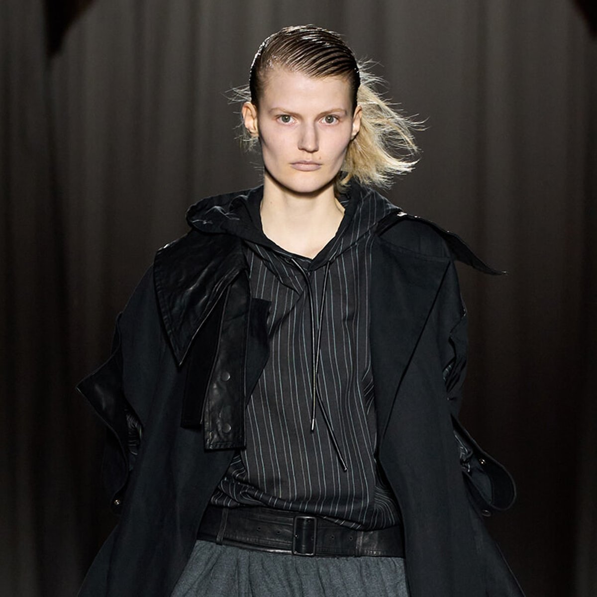 Runway model wearing a black hooded coat over a pinstripe shirt, with a gray belt and skirt, walking the catwalk.