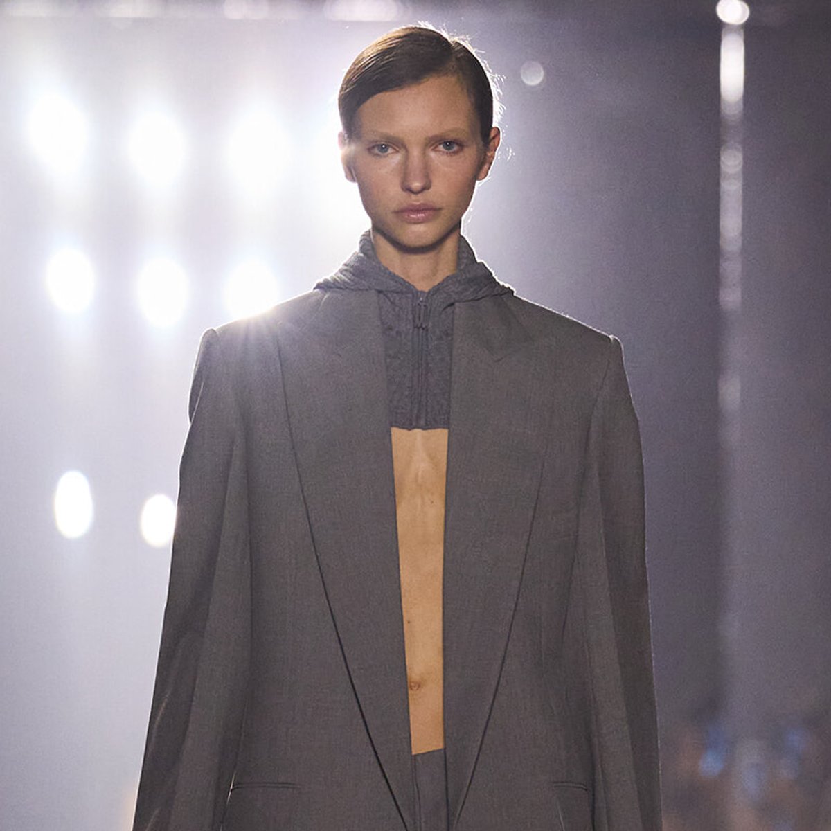 Model walks the runway in a gray oversized blazer with a dark hoodie underneath, bright backstage lights behind her.