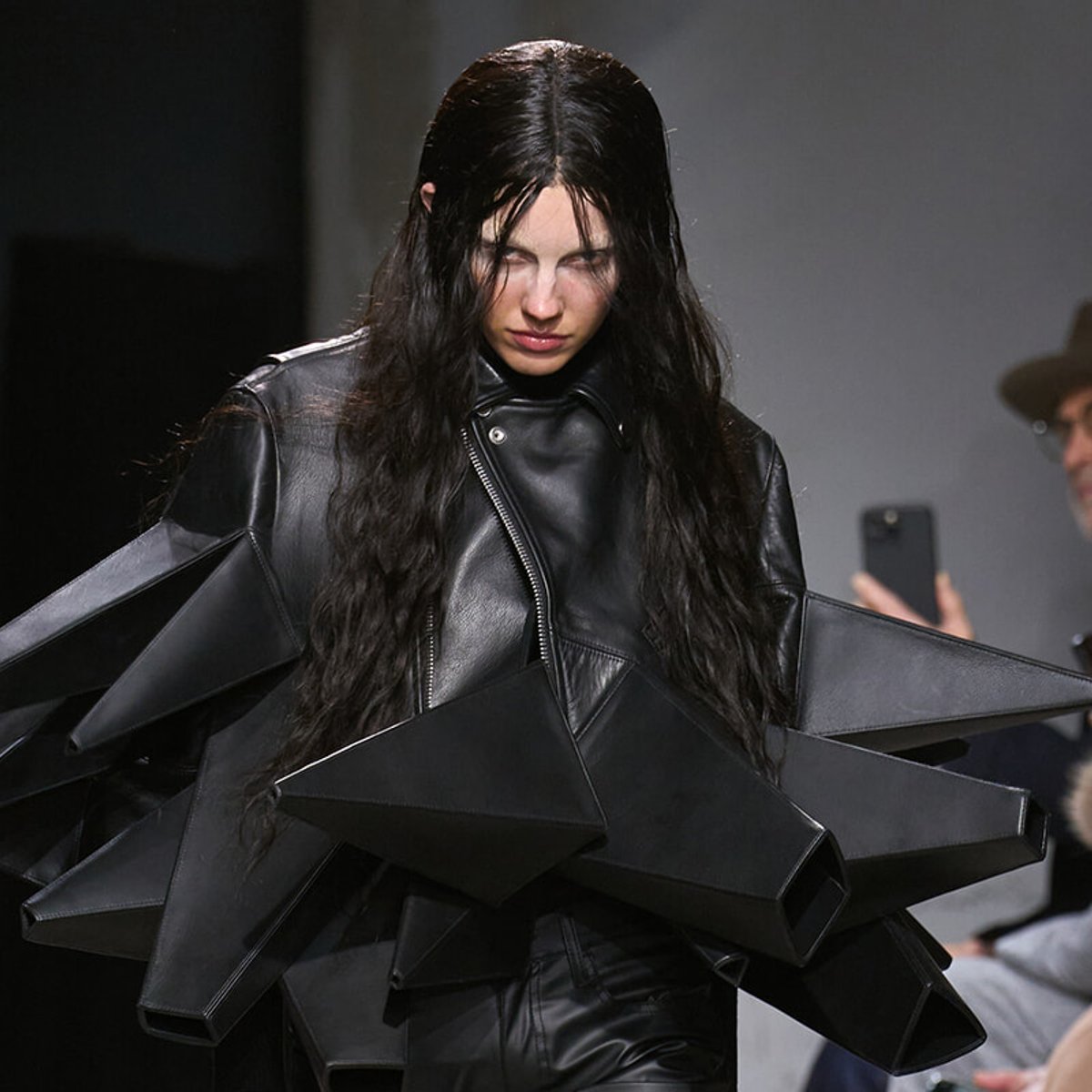 Runway model wearing a dramatic black leather outfit with oversized geometric angular panels in a fashion show setting.