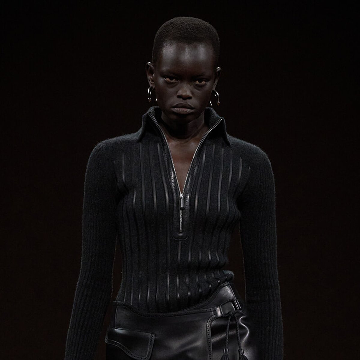 Model walking a dark runway in a black ribbed zip sweater and matching leather pants, minimalist styling.