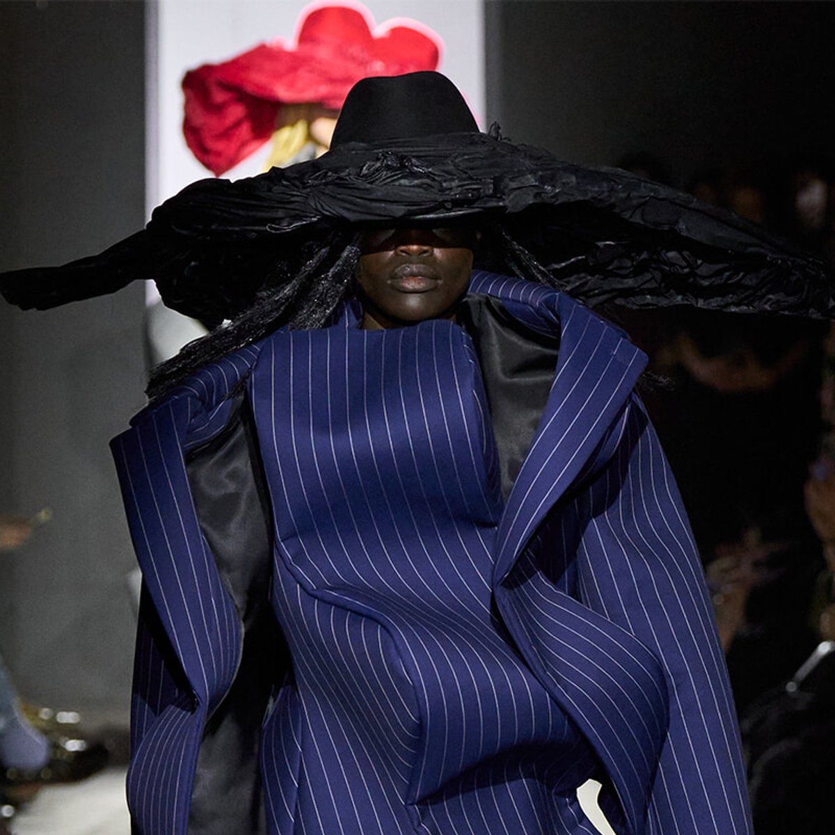 Model on runway wearing an oversized navy pinstripe suit with a broad black hat that covers the face.