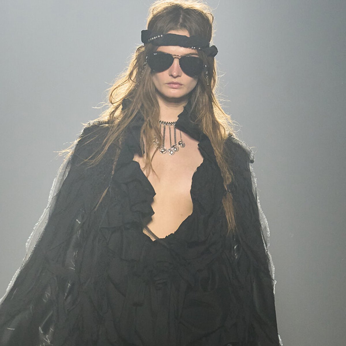 Model on a runway wearing a black, ruffled outfit with a headband, sunglasses, and layered necklace.