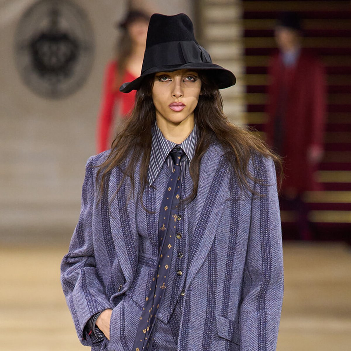 Runway model in a blue pinstripe suit with a striped shirt, tie, and a wide-brimmed black hat.