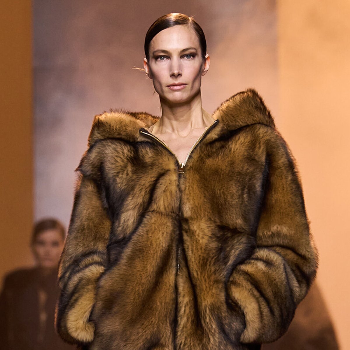 Model on a runway wearing a large brown fur coat with a front zipper, hands in pockets.