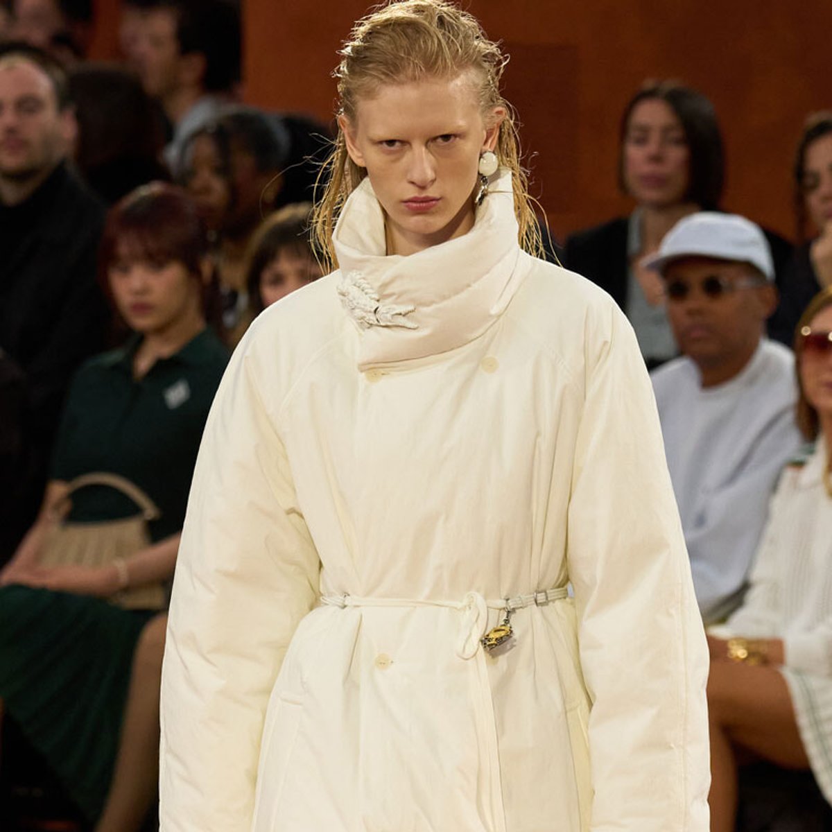 Model walking a fashion runway in a loose white coat with a high, wrapped collar and belt, audience in the background