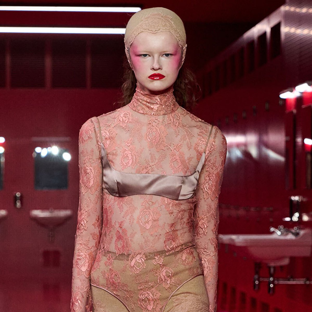 Model wearing a pink lace turtleneck dress with a satin bustier in a red-lit backstage setting, looking at the camera with bold makeup.