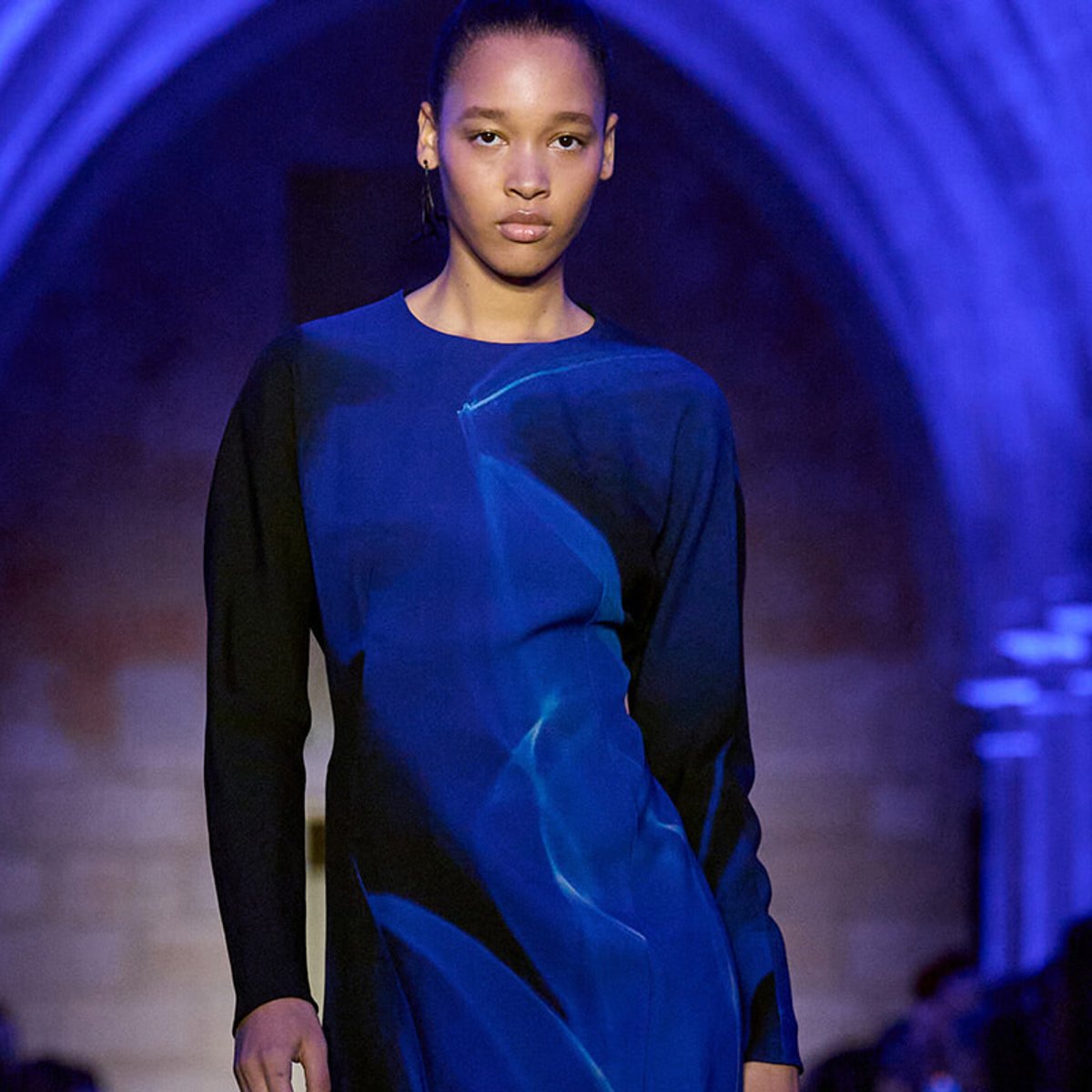 Female model walking a runway in a cobalt blue dress with black sleeves under blue lighting at a fashion show.
