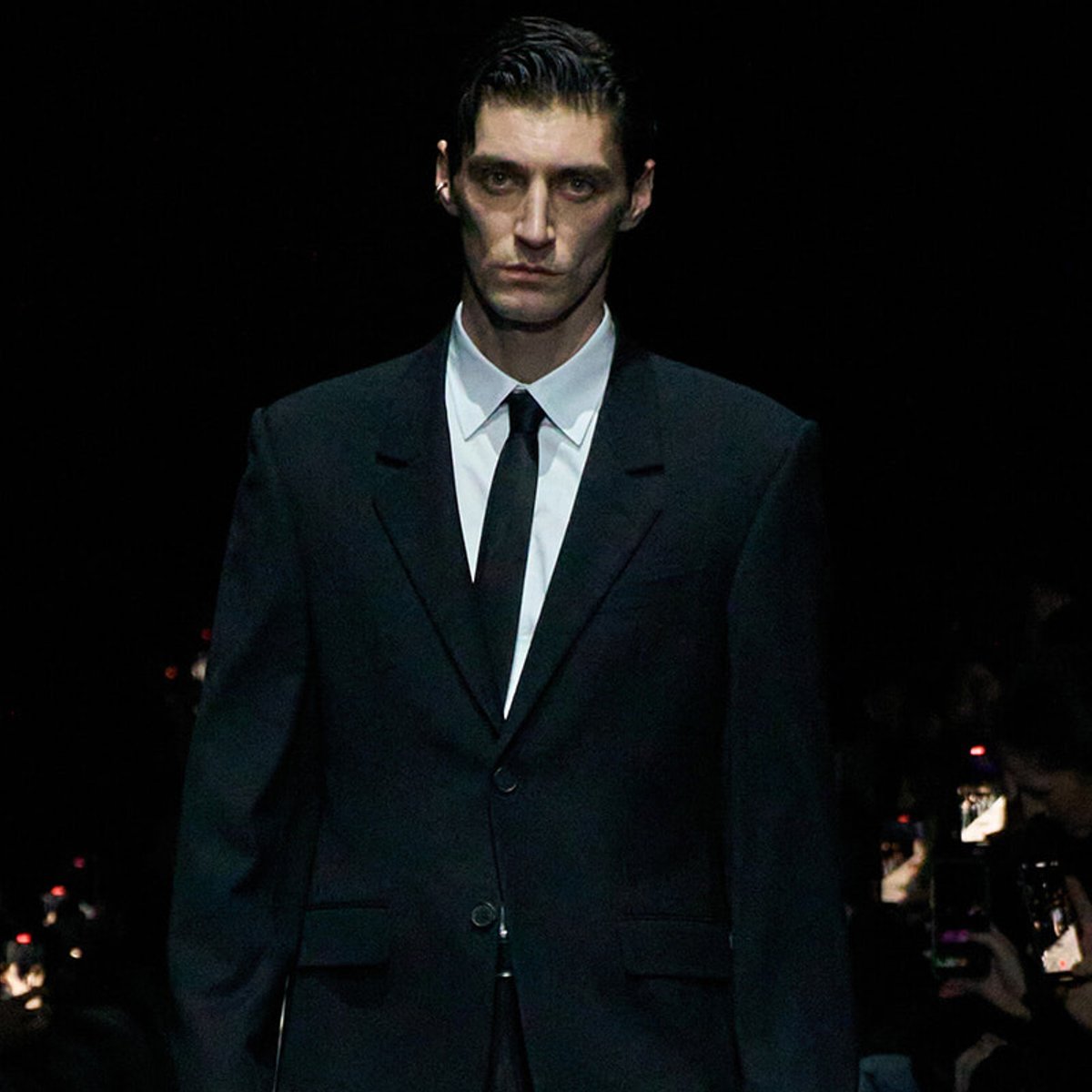 Male model in a black suit and black tie walking a runway against a dark background, white shirt visible underneath.