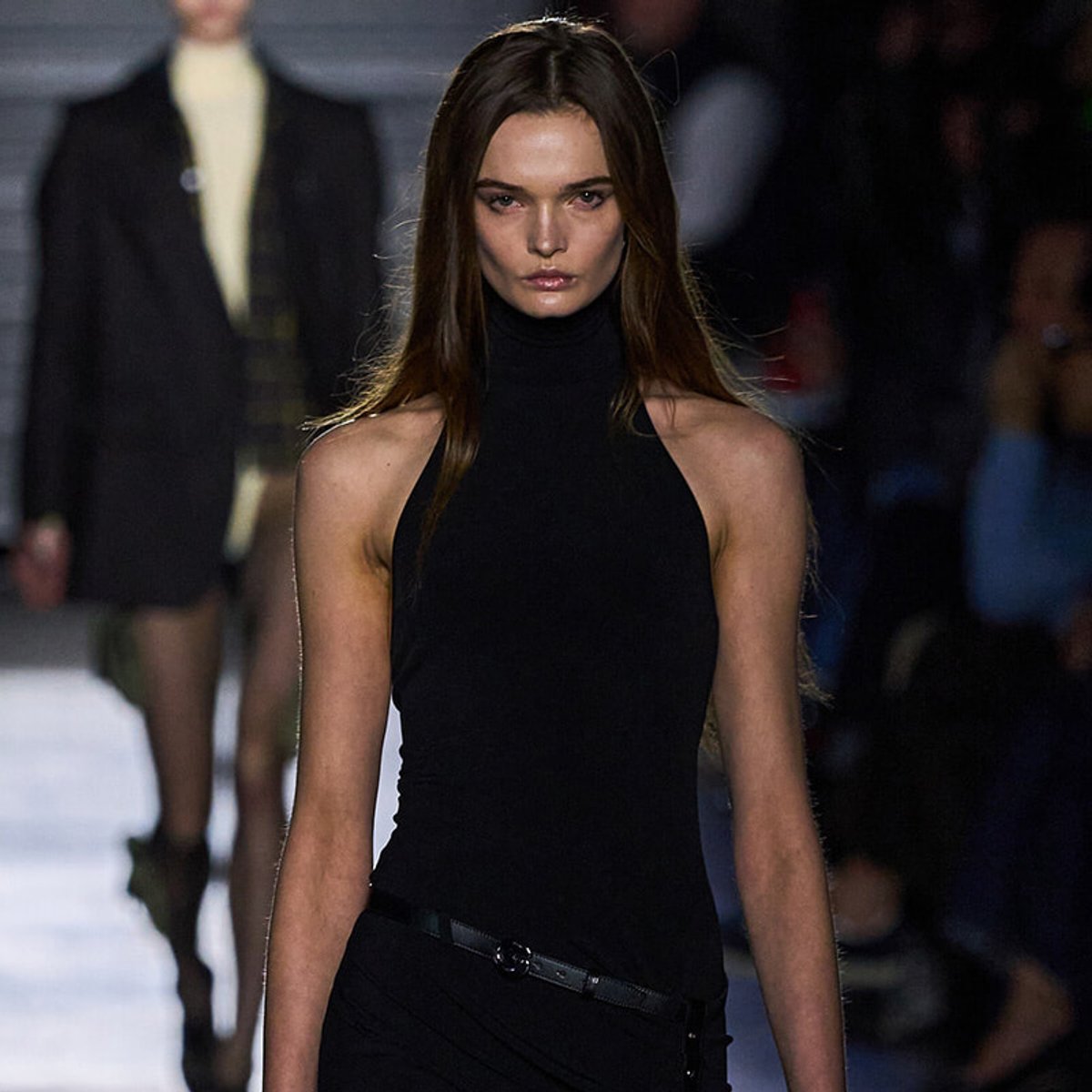 Model walking the runway in a fitted black sleeveless turtleneck top and belt, with a focused expression.