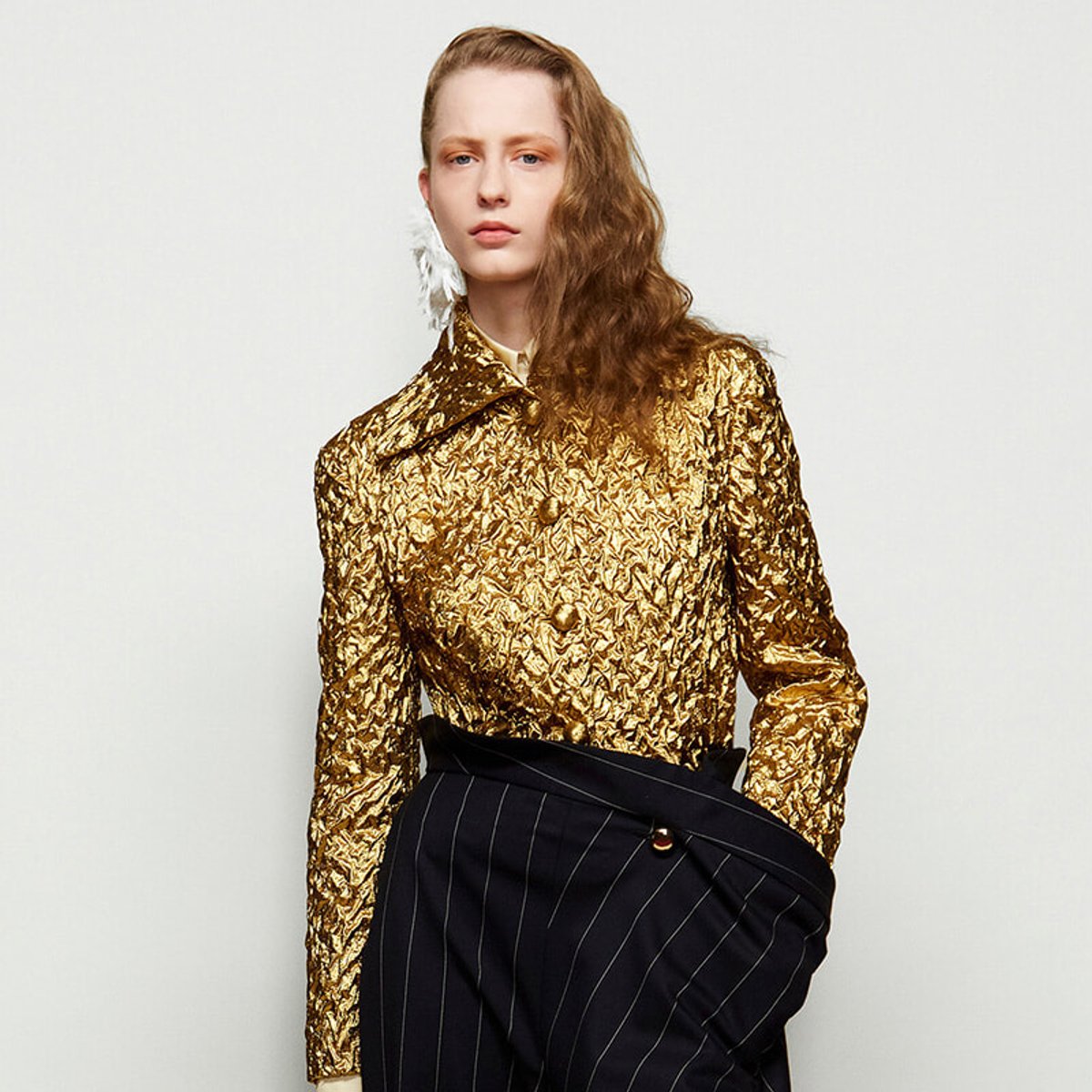Model wearing a gold metallic, crinkled blouse with a large collar and a navy pinstripe wrap skirt against a light background, fashion shoot