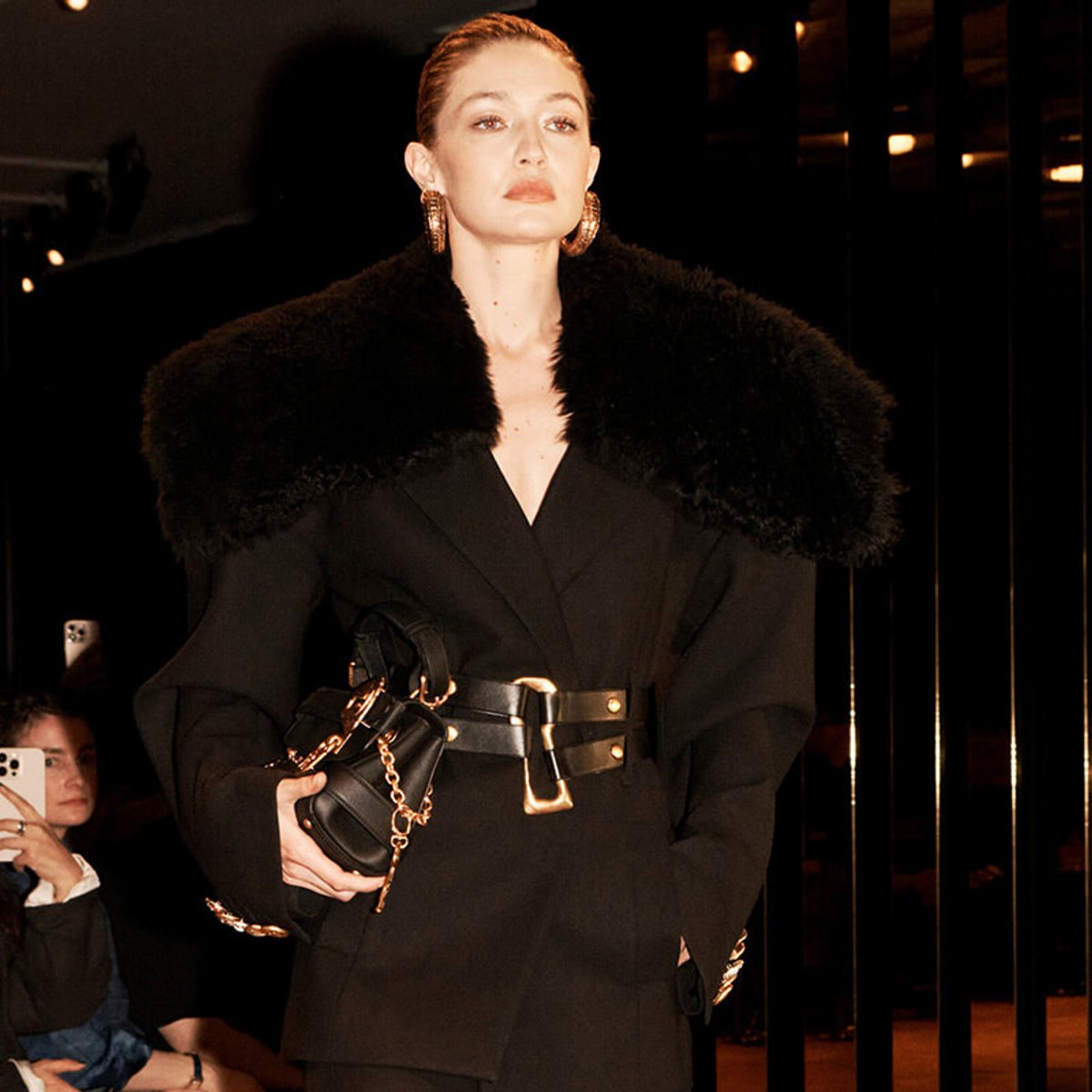 Female model on a runway wearing a black coat with a large fur collar, a wide gold belt, and a black handbag with a gold chain; audience members visible in the background.
