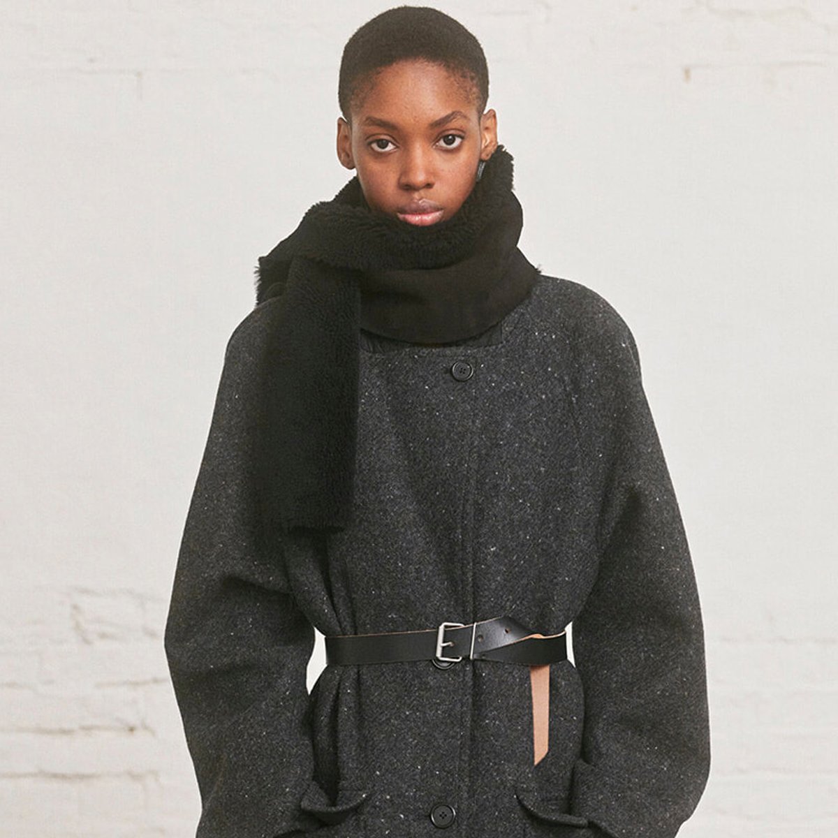 Person wearing a dark textured wool coat with a wide black belt and a black scarf, standing against a light background.