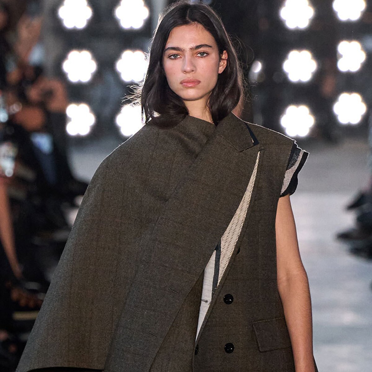 Model walks a fashion runway in a dark olive cape over a coat, bright stage lights in the background.