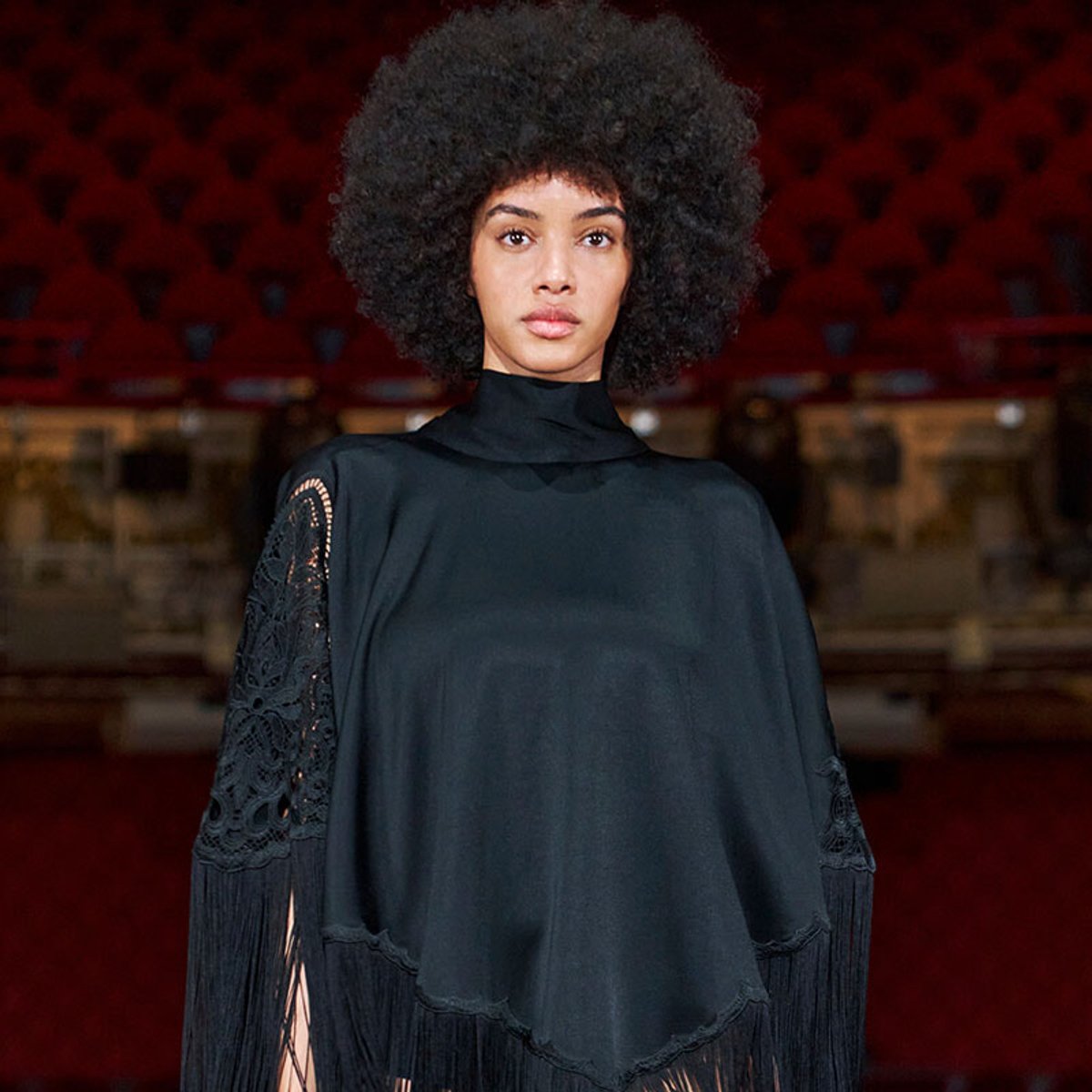 Woman with a voluminous afro wearing a black high-neck top with lace sleeves, standing in a theater with red seats in the background.