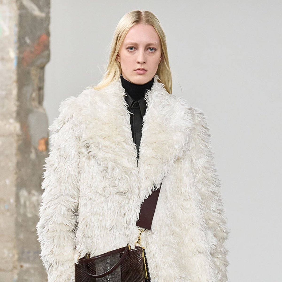Model wearing a plush white faux fur coat over a black turtleneck, carrying a brown textured handbag.