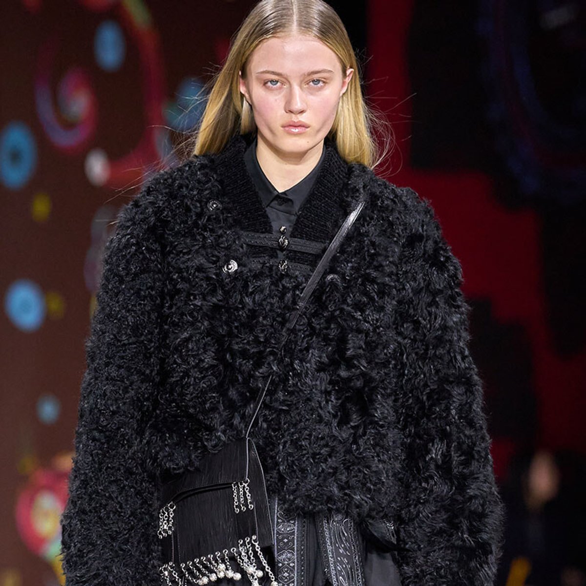 Model walks the runway in a black, fluffy textured coat over a dark outfit, carrying a small crossbody bag with chains.