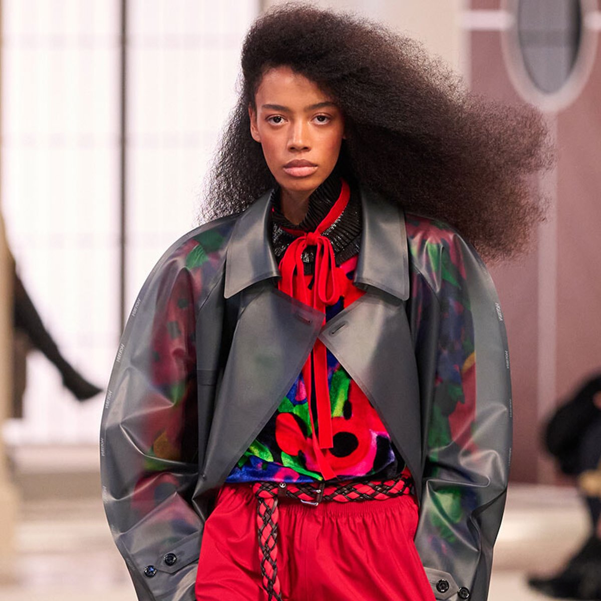 Model walking a fashion runway with voluminous natural hair, wearing a colorful top, red scarf, gray coat, and red drawstring shorts.