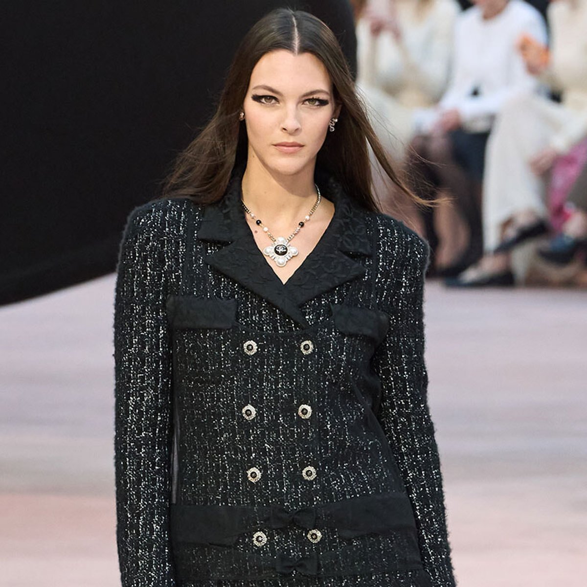 Model on a runway wearing a black textured double-breasted coat with silver-tone buttons and a statement necklace.