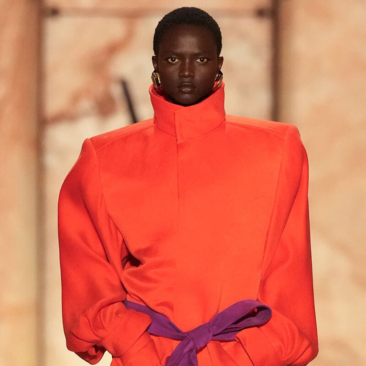 Fashion model walking the runway in a high-collar bright orange top with a purple belt tied at the waist, minimalist background.