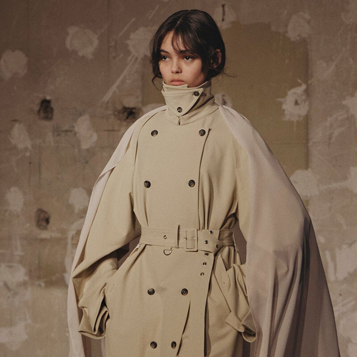 Female model wearing a beige double-breasted trench coat with a cape and high collar, standing against a textured wall.