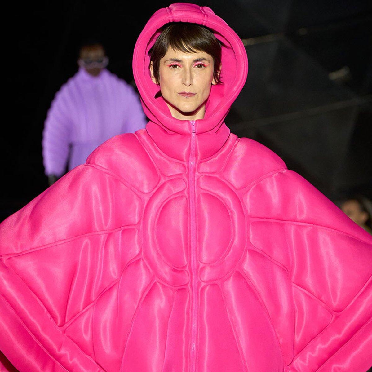 Person modeling a bright pink oversized puffer outfit with hood, resembling a cape on a runway.