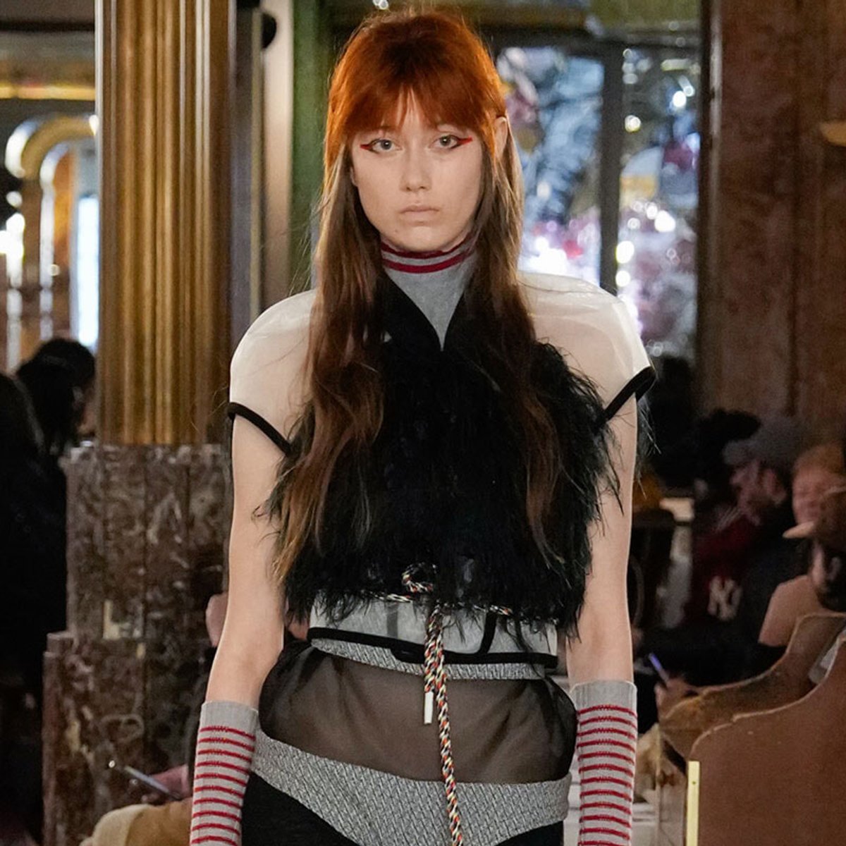 Model on a runway wearing an avant-garde outfit with a black feathered top, gray knit sleeves, and a metallic belt; red hair and bold makeup.