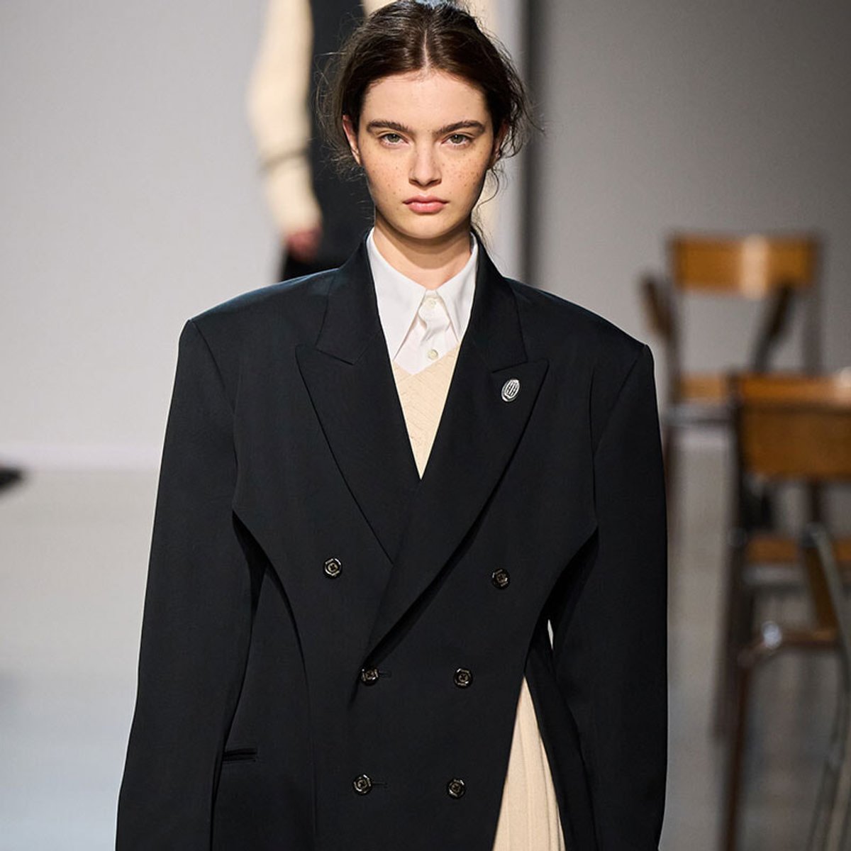 Runway model wearing a black oversized double-breasted blazer over a white shirt and beige knit, with a lapel pin, in a fashion-show setting.