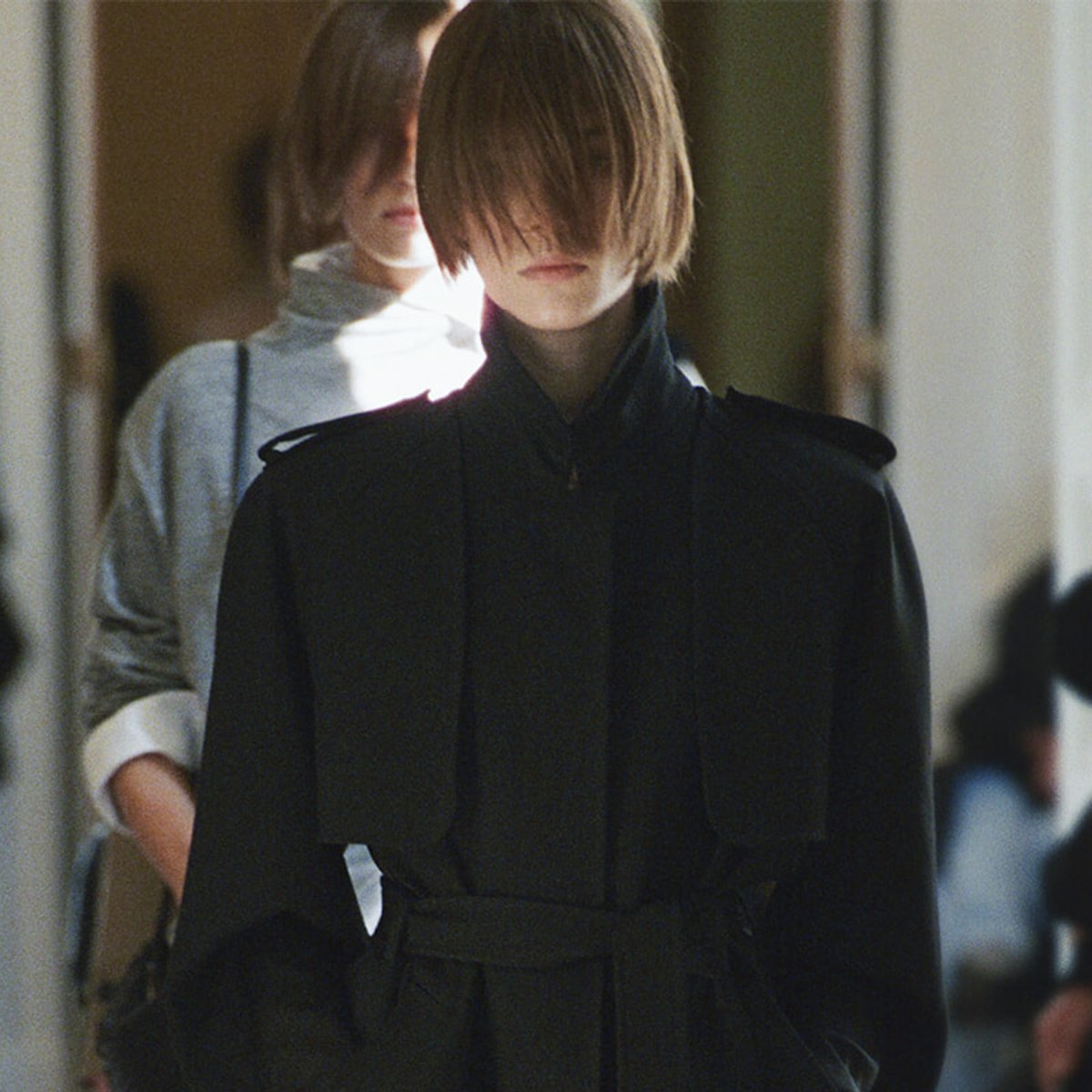Model wearing a dark belted trench coat with hair covering the face, walking in a hallway or backstage area; another person blurred behind.