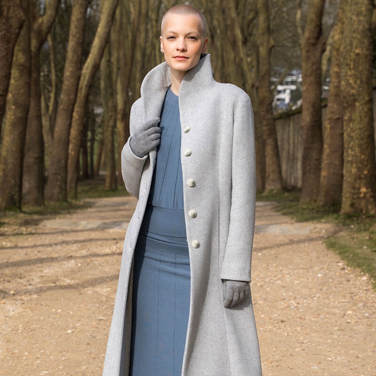Woman with a shaved head in a light gray, knee-length coat and blue dress, standing on a dirt path lined with trees in a park.