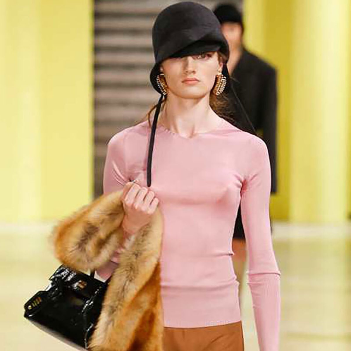 Fashion model walks a runway in a pale pink fitted long-sleeve top, a black hat, fur stole draped over her arm, and a glossy black handbag.