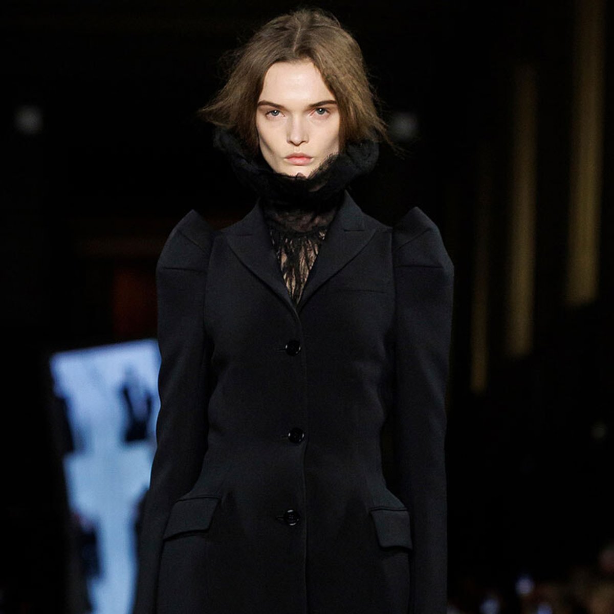 Female model walks the runway in a dark, tailored black coat with a high lace neck.