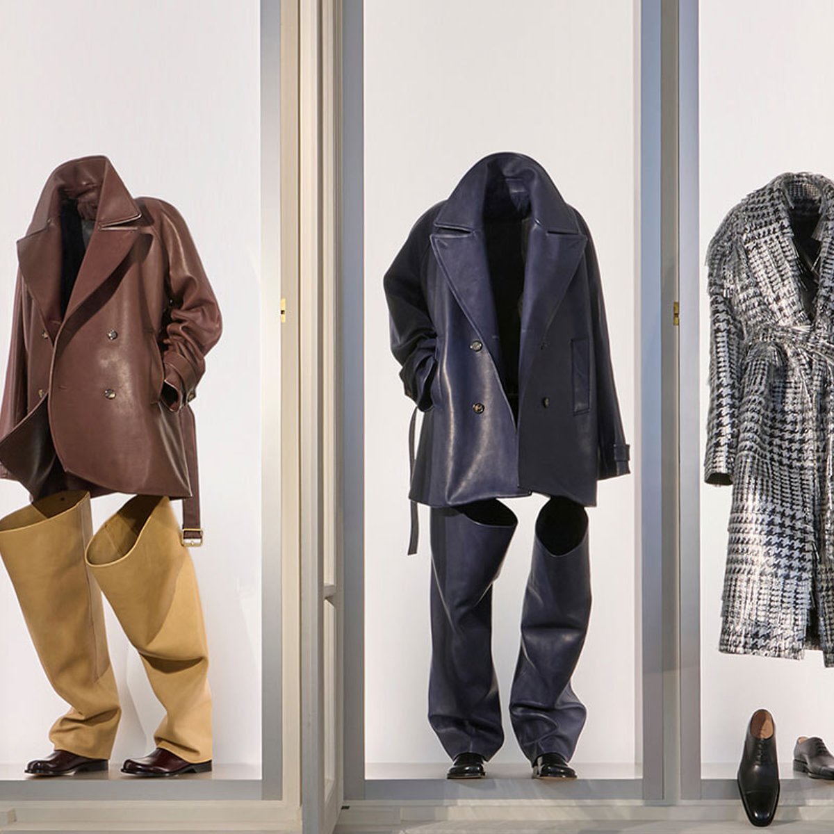 Window display featuring three mannequins in oversized coats: brown leather coat with tan wide-leg pants on the left, navy leather set in the center, and a black-and-white plaid coat on the right, with shoes visible.