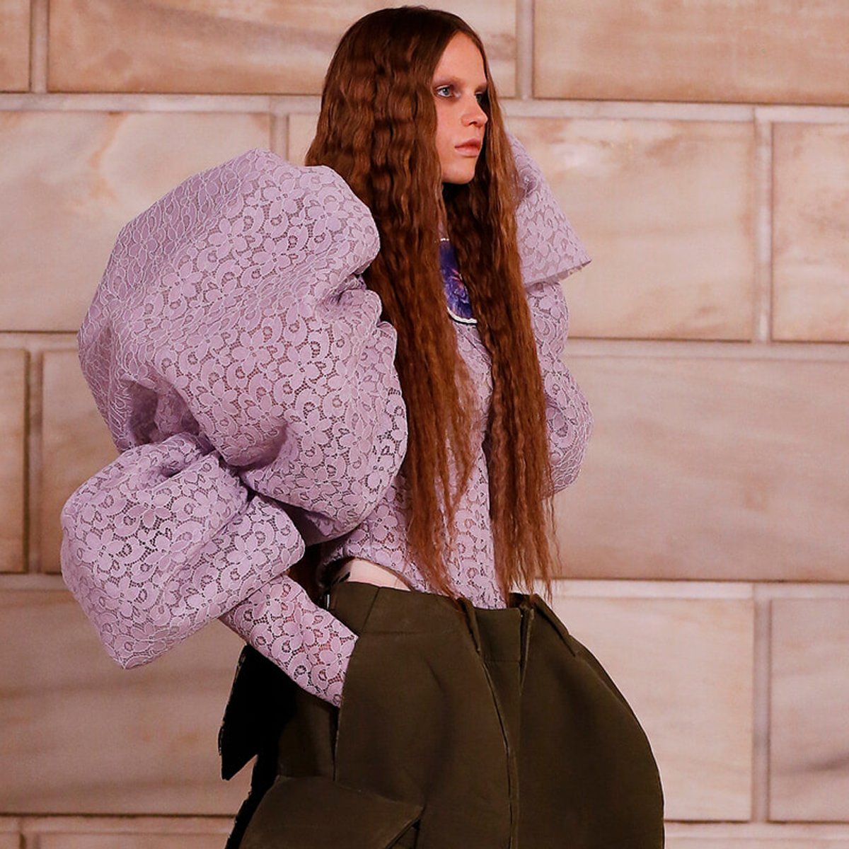 Model wearing a lavender lace top with huge puff sleeves and olive high-waisted pants, posing against a stone wall.