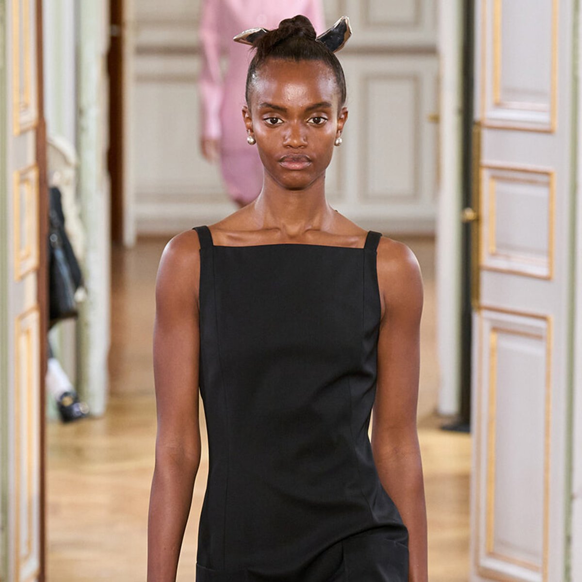Model wearing a black sleeveless dress walks down a hallway runway with a metallic bow headband in her hair.