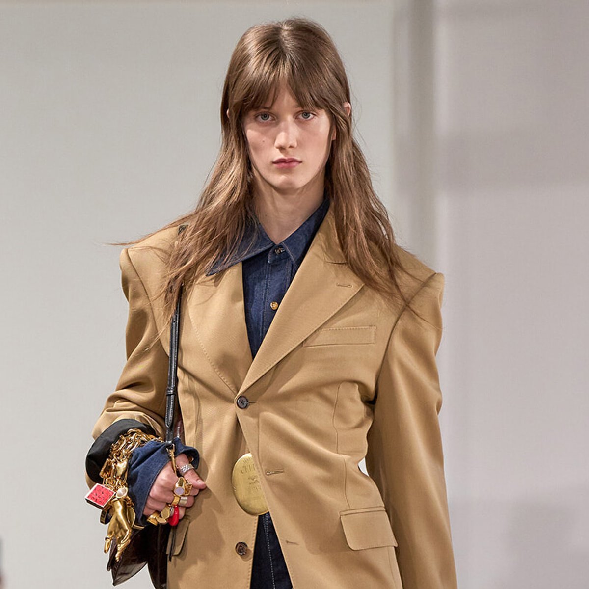 Model walking a runway wearing a tan blazer over a dark denim shirt, carrying a bag with accessories and keychains in her hand.