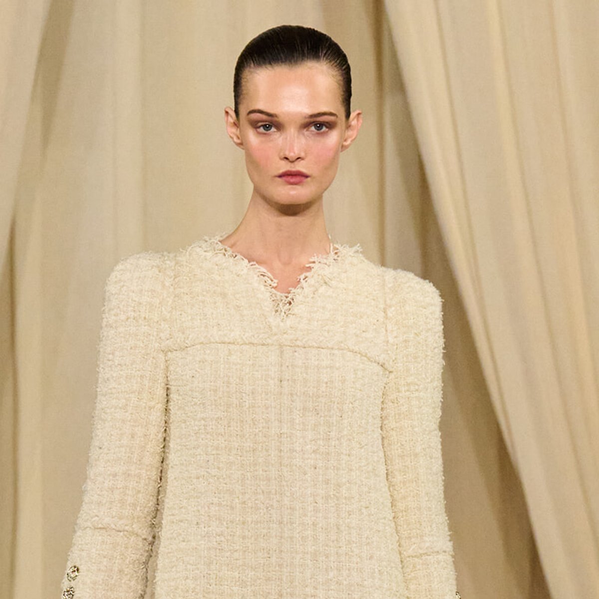 Model on a beige runway wearing a cream textured tweed dress with a high neckline and long sleeves.