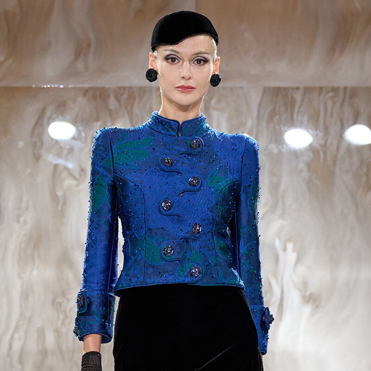 Model walking a runway wearing a royal blue, embellished military-style jacket with large decorative buttons and dark cuffs, paired with a black hat and earrings.