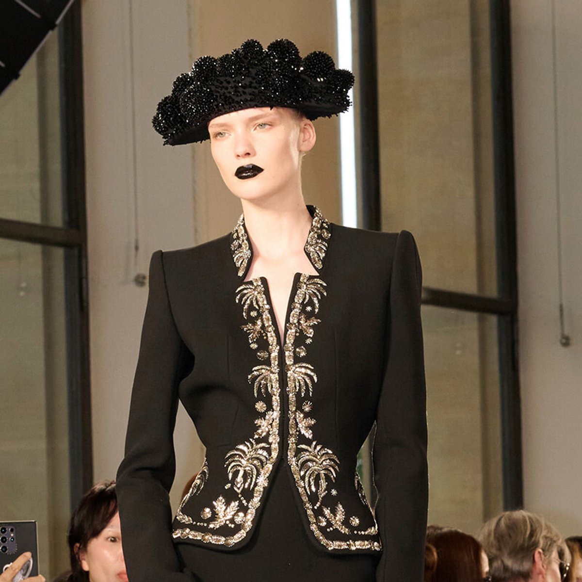 Model on a runway wearing a black tailored jacket with intricate silver embroidery and a large black beaded hat, eye-catching haute couture look.